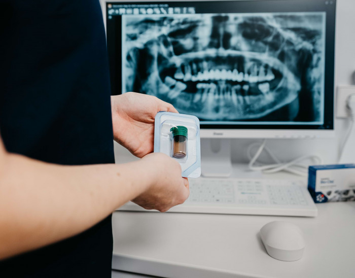 Eine Mitarbeiterin h&auml;lt ein Pr&auml;parat f&uuml;r Knochenaufbau vor einen Bildschirm mit einer R&ouml;ntgenaufnahme darauf.