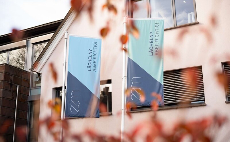 Nahaufnahme von Flaggen mit dem Logo vom Zahn&auml;rztezentrum Markgr&auml;flerland sowie der Aufschrift "L&auml;cheln? ABER RICHTIG!".