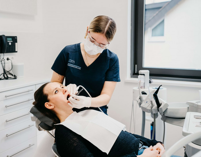 Eine Mitarbeiterin behandelt eine Patientin, die mit offenem Mund auf einem Behandlungsstuhl sitzt.