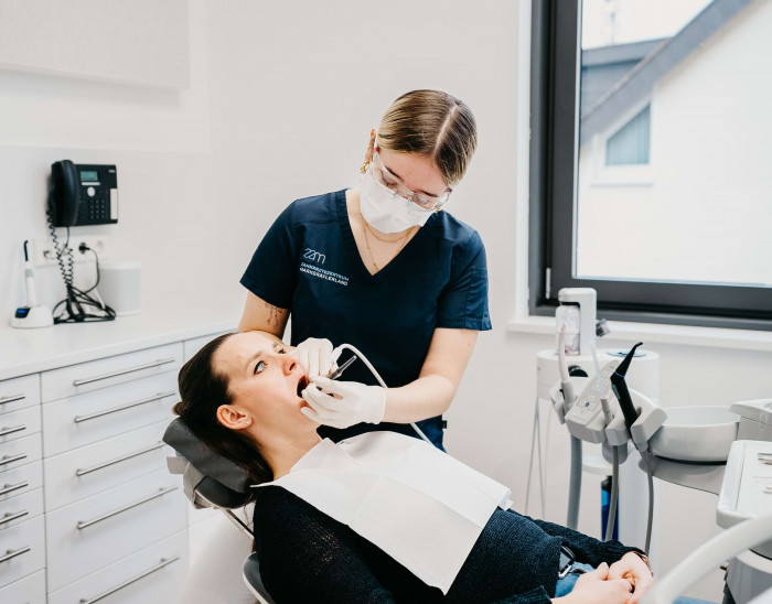 Eine Mitarbeiterin behandelt eine Patientin, die mit offenem Mund auf einem Behandlungsstuhl sitzt.