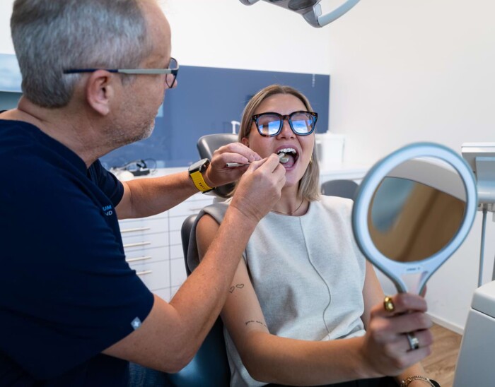 Dr. Brunner behandelt eine Patientin. Die Patientin h&auml;lt sich einen Handspiegel vor das Gesicht.