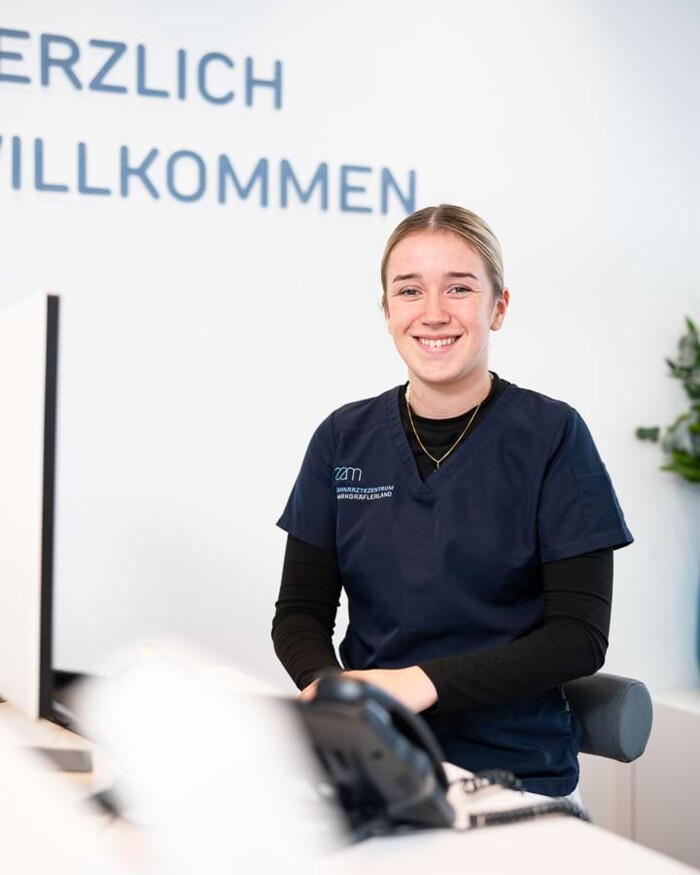 Eine Auszubildende sitzt am Empfang und l&auml;chelt in die Kamera.